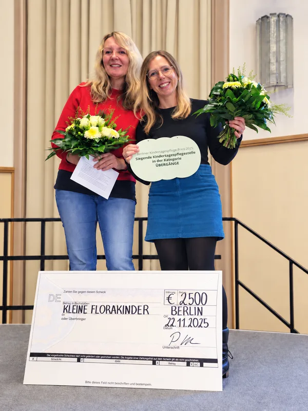 Christine Zekina und Sandra Ebeling mit Blumensträußen und dem Preisschild „Siegende Kindertagespflegestelle in der Kategorie Übergänge“, vor ihnen der Scheck über 2.500 Euro